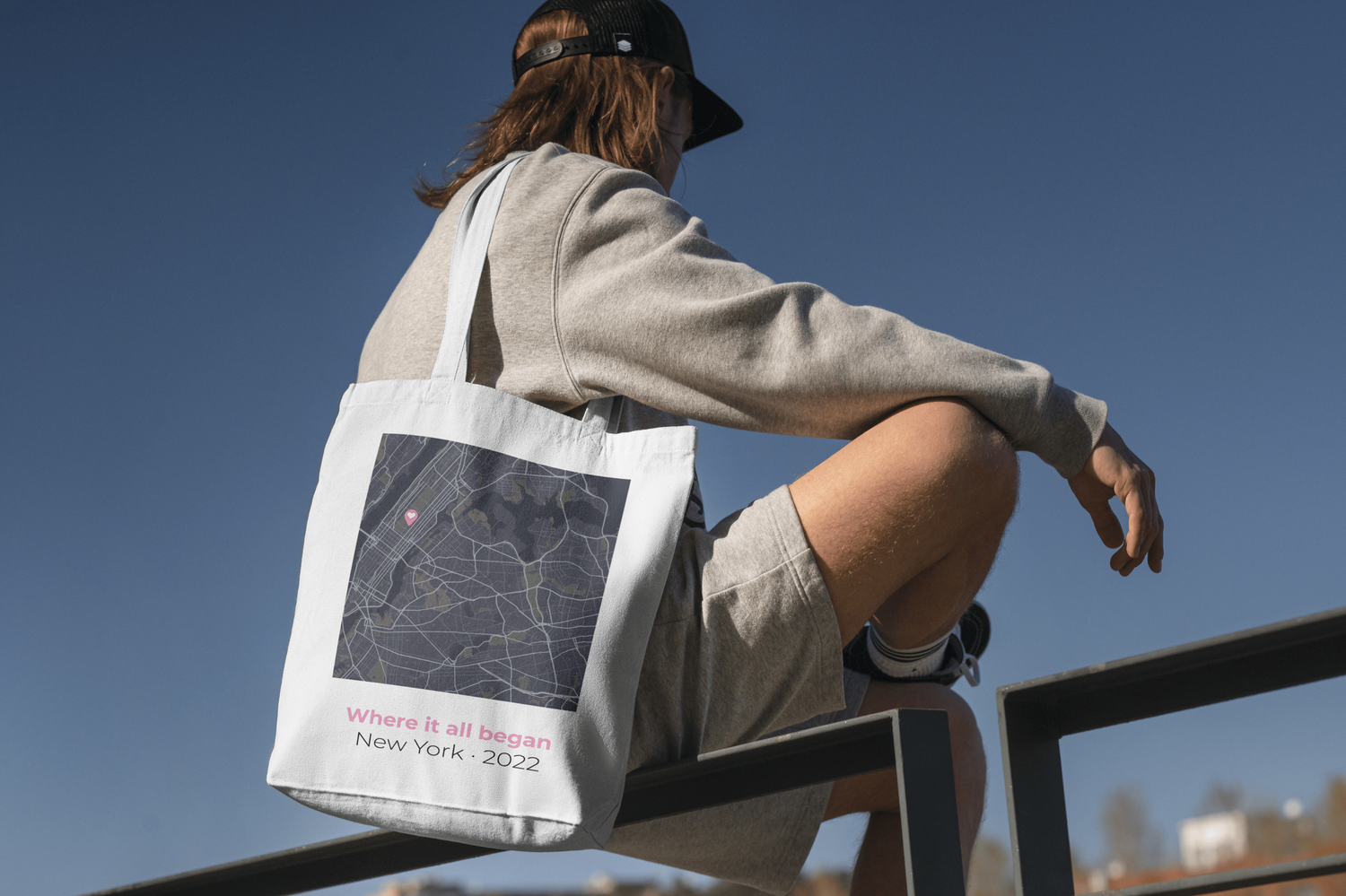 Person sitting on a railing with a tote bag featuring a map design against a clear blue sky.